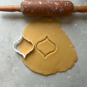 May include: A wooden rolling pin dusted with flour is positioned above a sheet of dough. A white plastic cookie cutter in a decorative shape sits next to the cut-out dough. Baking and pastry concept.