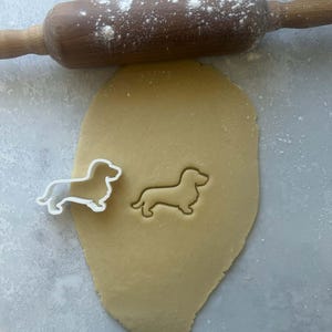 May include: A wooden rolling pin dusted with flour rests above a sheet of dough. A dachshund-shaped cookie cutter sits next to an imprint of the same shape in the dough. Baking and cookie-making concept.