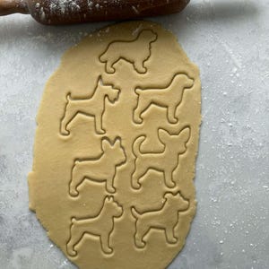 May include: A wooden rolling pin rests above a sheet of dough with dog-shaped cookie cutouts. The dough is light brown, and the cutouts are in various dog breeds. Flour dusts the surface, suggesting baking.