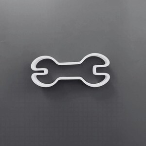 May include: A white plastic cookie cutter shaped like a double-ended open-ended wrench. The tool features a central handle connecting two circular heads, each with a U-shaped opening at the ends, designed for cutting dough into a wrench shape.
