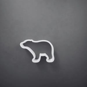 Pode incluir: Um cortador de biscoitos branco em forma de urso com centro oco e contorno definido. O objeto apresenta a silhueta distinta de um urso a caminhar com cabeça arredondada, costas curvas e quatro patas, concebido para cortar formas.