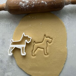 May include: A wooden rolling pin dusted with flour rests above a sheet of dough. A white plastic dog-shaped cookie cutter sits next to an imprint of the same shape in the dough. The dough is light brown.