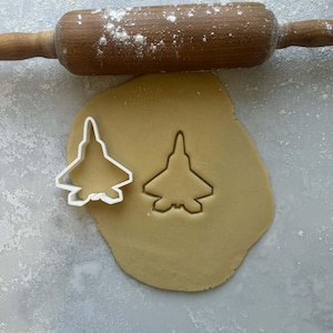 Puede incluir: Un cortador de galletas blanco en forma de avión junto a un corte en masa extendida. Un rodillo de madera espolvoreado con harina descansa sobre la masa. La escena se desarrolla sobre una superficie gris clara.