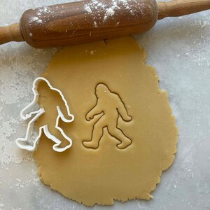 Pode incluir: Um rolo de massa de madeira repousa sobre uma folha de massa com um cortador de biscoitos Bigfoot e uma impressão. A massa é marrom clara e o cortador é branco.