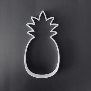 Op de afbeelding: Een witte koekjesvorm in de vorm van een ananas met een duidelijke omtrek, voorzien van een afgeronde basis en een puntig, meerpuntig bovendeel dat de bladeren voorstelt. Het object is opgebouwd met een dun, uniform frame.