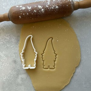 Op de afbeelding: Een houten deegroller bestrooid met bloem ligt boven een vel deeg. Een witte gnome-vormige koekjesvorm staat naast een afdruk van dezelfde vorm in het deeg. Het deeg is lichtbruin en de achtergrond is lichtgrijs.