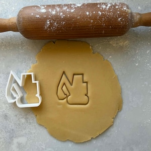 May include: A wooden rolling pin dusted with flour rests above a circle of tan cookie dough. A white plastic cookie cutter in the shape of a construction vehicle sits next to the cut-out shape in the dough. Baking and construction themed.
