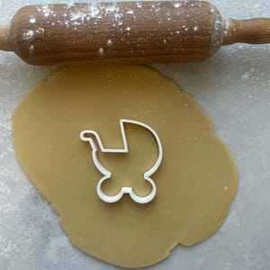 Op de afbeelding: Een witte plastic babywagenkoekjesvorm op een deegvel, met een houten deegroller bestrooid met bloem op de achtergrond. Het deeg is lichtbruin en de deegroller is bruin.