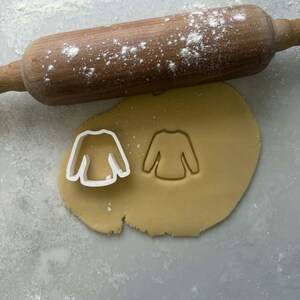 Könnte beinhalten: Ein mit Mehl bestäubter Nudelholz aus Holz liegt über einem Teigblatt. Ein weißer Kunststoff-Ausstecher in Pulloverform liegt auf dem Teig neben einem Pulloverförmigen Abdruck. Der Teig ist hellbraun.