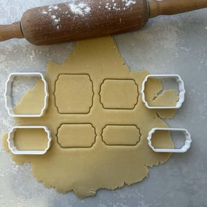 Puede incluir: Un rodillo de madera espolvoreado con harina descansa sobre una lámina de masa con seis cortadores de galletas blancos. Los cortadores tienen varias formas rectangulares, algunas con bordes decorativos, y han cortado la masa.