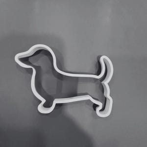 May include: A white cookie cutter shaped like a dachshund dog in profile. The outline features the distinct long body, short legs, floppy ears, and an upward-curving tail of the breed. The cutter has a smooth, uniform edge and a hollow center.