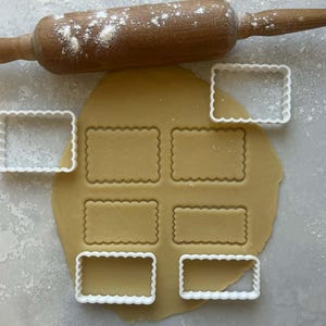 May include: A wooden rolling pin dusted with flour rests above a sheet of dough with rectangular cutouts. White, scalloped-edge cookie cutters surround the dough, ready for baking. The scene suggests a home baking activity.