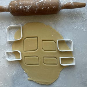 Op de afbeelding: Een houten deegroller bestrooid met bloem ligt boven een deegvel met koekjesvormen. Witte plastic koekjessnijders zijn rond het deeg geplaatst, dat is uitgesneden in afgeronde rechthoekige vormen.