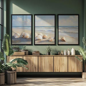 May include: Three framed beach scene paintings. Each features a sandy shore with seashells and ocean waves under a cloudy sky. The artwork is displayed above a wooden cabinet with plants.