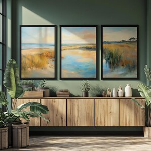 Op de afbeelding: Drie ingelijste landschapsschilderijen met zwarte frames hangen boven een houten kast. De schilderijen tonen kusttaferelen met zandstranden, water en moerasgrassen in tinten geel, blauw en oranje. Decoratieve planten en vazen staan op de kast.