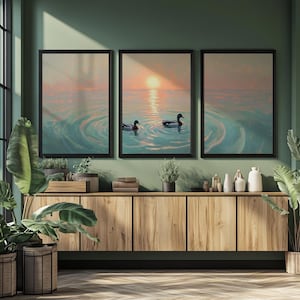 May include: Three framed landscape paintings depict a serene sunset over water, with ducks swimming in the rippling water. The artwork features a color palette of soft blues, pinks, and oranges, creating a calming atmosphere. The frames are black.