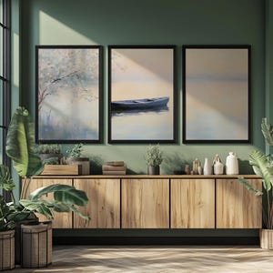 Könnte beinhalten: Drei gerahmte Landschaftsbilder mit schwarzen Rahmen hängen über einem Holzschrank. Die Gemälde zeigen einen Baum, ein Boot auf dem Wasser und eine ruhige Wasserszene. Der Raum hat grüne Wände und ist mit Pflanzen dekoriert.