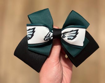 May include: A multi-layered hair bow featuring dark green, black, and white grosgrain ribbon. The centre of the bow displays a white ribbon section printed with a repeating black and teal eagle logo. The layers are secured with a black ribbon knot.