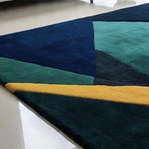 May include: A geometric area rug featuring a modern design. The rug showcases a patchwork of shapes in shades of navy, teal, and green, with a bold yellow stripe. The rug's texture appears soft and plush, adding visual interest to the floor covering.
