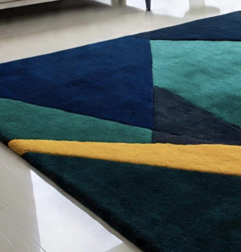 May include: A geometric area rug featuring a modern design. The rug showcases a patchwork of shapes in shades of navy, teal, and green, with a bold yellow stripe. The rug's texture appears soft and plush, adding visual interest to the floor covering.
