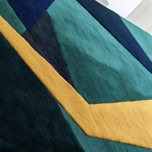 May include: Geometric patterned rug featuring a combination of teal, navy, and yellow. The design incorporates angular shapes and a textured surface, creating a modern and artistic aesthetic. The rug's color palette adds a vibrant touch to any room.
