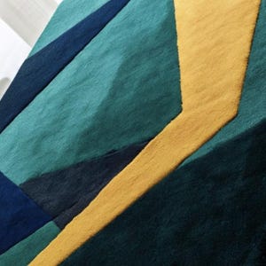 May include: A geometric area rug featuring a bold design with teal, navy, and gold sections. The rug's pattern includes angular shapes and a textured surface, adding visual interest to any room. The rug is a modern home decor item.
