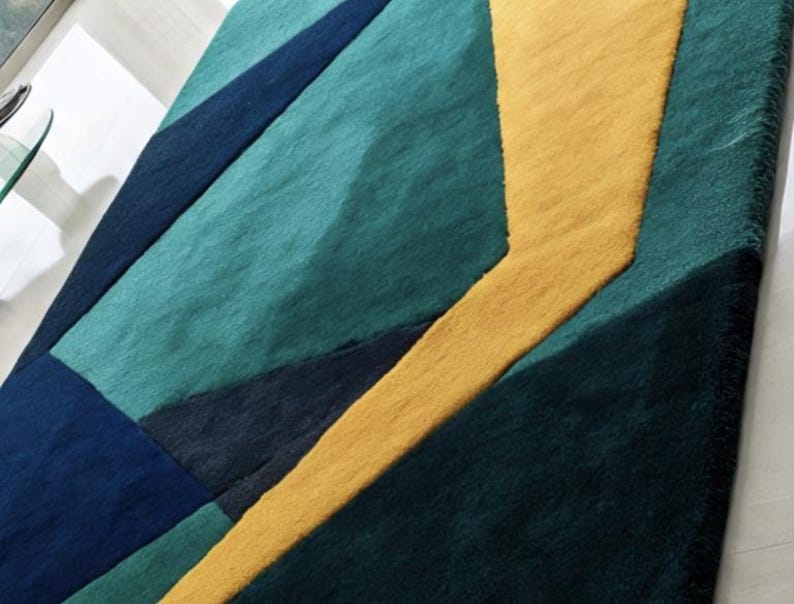 May include: A geometric area rug featuring a bold design with teal, navy, and gold sections. The rug's pattern includes angular shapes and a textured surface, adding visual interest to any room. The rug is a modern home decor item.