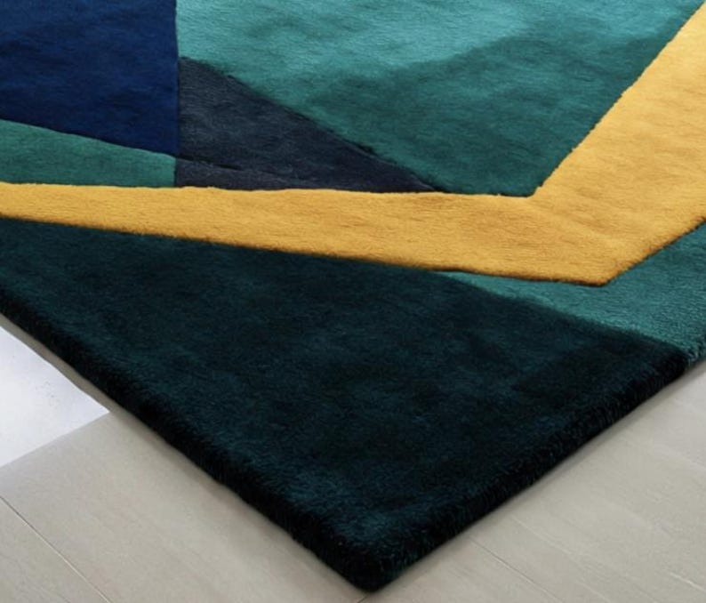 May include: A geometric area rug featuring a modern design with a combination of teal, navy, and gold colors. The rug's pattern includes angular shapes and lines, creating a contemporary aesthetic. The rug is made of a soft, plush material.