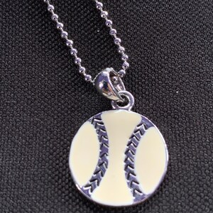 Könnte beinhalten: Halskette mit Baseball-Anhänger aus Silber. Der Anhänger hat eine runde Form mit cremefarbenem Hintergrund und dunkelblauen Nahtdetails. Die Halskette hat eine silberne Kugelkette.
