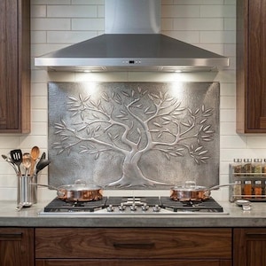 May include: Kitchen interior with a stainless steel range hood above a gas cooktop. A decorative metal tree design backsplash is centered above the cooktop. Copper-bottomed pans sit on the stove. Dark wood cabinets are on either side.