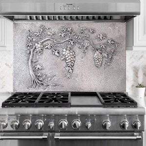 May include: A stainless steel kitchen backsplash with a detailed relief of a grape vine. The design features a tree trunk, leaves, and clusters of grapes. The backsplash is mounted above a stainless steel gas range.