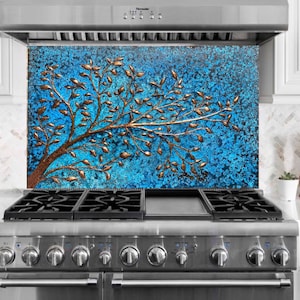 May include: A blue and copper-colored kitchen backsplash featuring a tree design. The tree has copper-colored leaves and branches against a textured blue background. The backsplash is installed above a stainless steel gas range.