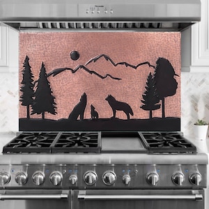 Puede incluir: Una placa para salpicaduras de campana extractora de color cobre con un diseño de silueta negra de una escena de montaña con árboles, lobos y una luna llena. La placa para salpicaduras está montada sobre una cocina de gas de acero inoxidable.