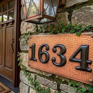 以下が含まれることがあります： 黒い数字「1634」が刻まれた銅色の住所プレート。石壁に取り付けられ、緑のツタに囲まれています。木製のドアとランタンが背景に見えます。