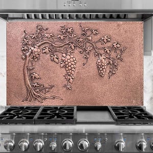 May include: A copper-colored kitchen backsplash with a detailed relief carving of a grape vine. The design features a tree trunk, branches, leaves, and clusters of grapes. The backsplash is installed above a stainless steel gas range.