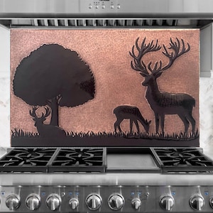 May include: A copper-colored range hood backsplash with a black silhouette of a tree, deer, and grass. The backsplash is installed above a stainless steel gas range with black grates.