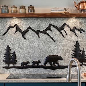 May include: A decorative kitchen backsplash featuring a black mountain range, pine trees, and a bear family silhouette. The artwork is mounted on a textured, light-colored background, creating a rustic, nature-inspired theme.