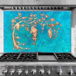 May include: A copper-colored vine design with grapes is set against a turquoise background. The artwork is mounted above a stainless steel gas range. The vine and grape design is a decorative element for the kitchen.