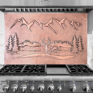 Könnte beinhalten: Kupferfarbene Dunstabzugshauben-Rückwand mit einer Berg- und Waldszene. Das Design zeigt einen Hirsch, Bäume und Berge. Das Kupfer hat eine strukturierte Oberfläche und ist über einem Gasherd aus Edelstahl montiert.