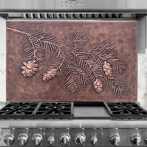 May include: A copper range hood splashback with an embossed design of a pine branch and pine cones. The rectangular splashback is mounted above a stainless steel gas range. The copper has a warm, reddish-brown hue.