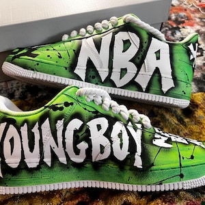 May include: Pair of custom sneakers with a green and black splatter design. The shoes feature the words "NBA" and "YOUNG BOY" in white graffiti-style lettering. White laces and soles complete the look.