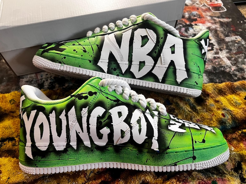 May include: Pair of custom sneakers with a green and black splatter design. The shoes feature the words "NBA" and "YOUNG BOY" in white graffiti-style lettering. White laces and soles complete the look.