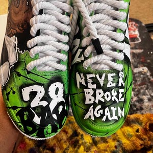 May include: Customized white sneakers with green and black accents. One shoe features the number "28" in white, while the other reads "NEVER BROKE AGAIN" in white. The shoes have white laces.