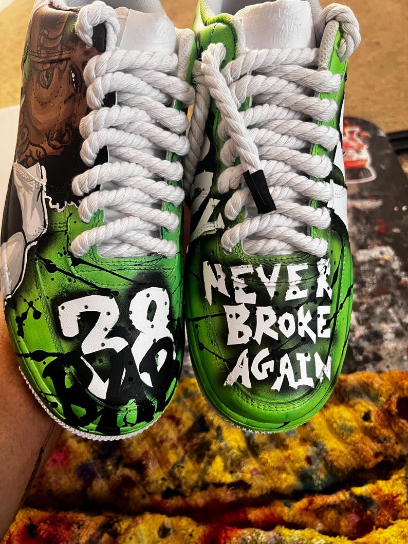 May include: Customized white sneakers with green and black accents. One shoe features the number "28" in white, while the other reads "NEVER BROKE AGAIN" in white. The shoes have white laces.