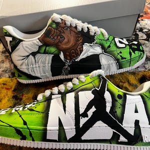 May include: Custom-painted sneakers with a vibrant green and black design. One shoe features a portrait, the other displays "NBA" and a silhouette. White laces and soles complete the look. A unique, artistic footwear item.