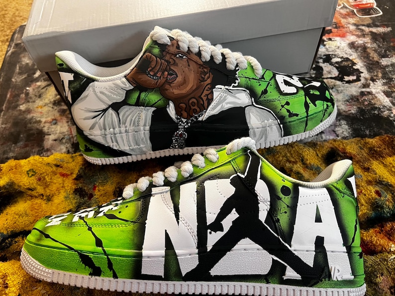 May include: Custom-painted sneakers with a vibrant green and black design. One shoe features a portrait, the other displays "NBA" and a silhouette. White laces and soles complete the look. A unique, artistic footwear item.