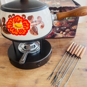 May include: A vintage fondue set with a speckled white pot decorated with an orange, red, and brown floral pattern. The set includes a black base, wooden-handled forks, and a box with "Fondue Set".