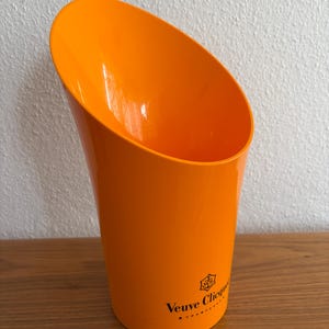 Może przedstawiać: Jasnopomarańczowe wiadro na szampana z ukośną górą. Logo Veuve Clicquot jest wydrukowane na boku na czarno. Wiadro wykonane jest z błyszczącego tworzywa sztucznego i stoi na drewnianej powierzchni.