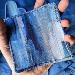 May include: A square, blue fabric coaster with a watercolor-like design in shades of blue and white. The coaster has a stitched border and is held in a hand. Another coaster with a heart shape is visible in the background.