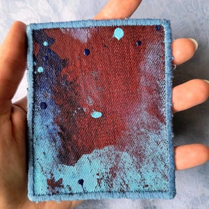 Może przedstawiać: Kwadratowa naszywka z niebieską obwódką i abstrakcyjnym wzorem. Naszywka przedstawia mieszankę czerwonych i niebieskich plam farby, z dodatkowymi niebieskimi kroplami farby rozrzuconymi po powierzchni. Krawędzie są starannie obszyte niebieską nicią.